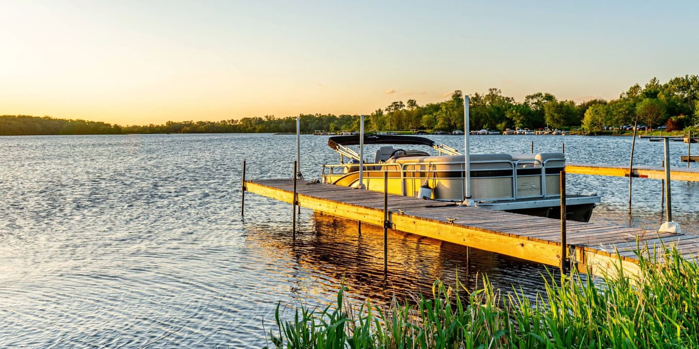 A pontoon boat protected by Iowa boat insurance docked at a wooden pier on a lake at sunset