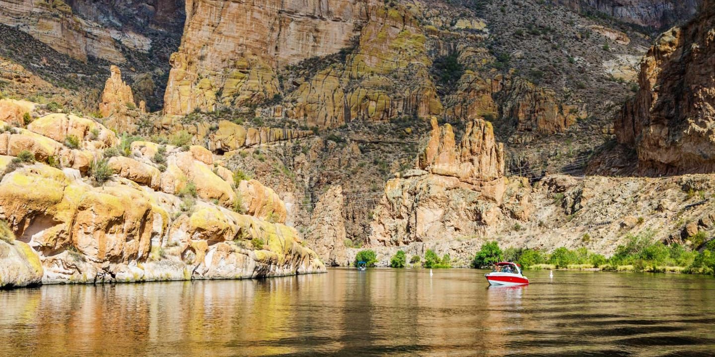 A small red and white motorboat, likely covered by Arizona boat insurance, floats on a calm river flowing through a deep canyon with rocky, weathered cliffs.