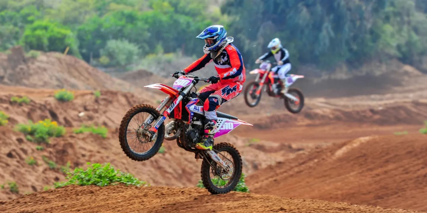 Two dirt bike riders jumping over mounds on an off-road motocross track