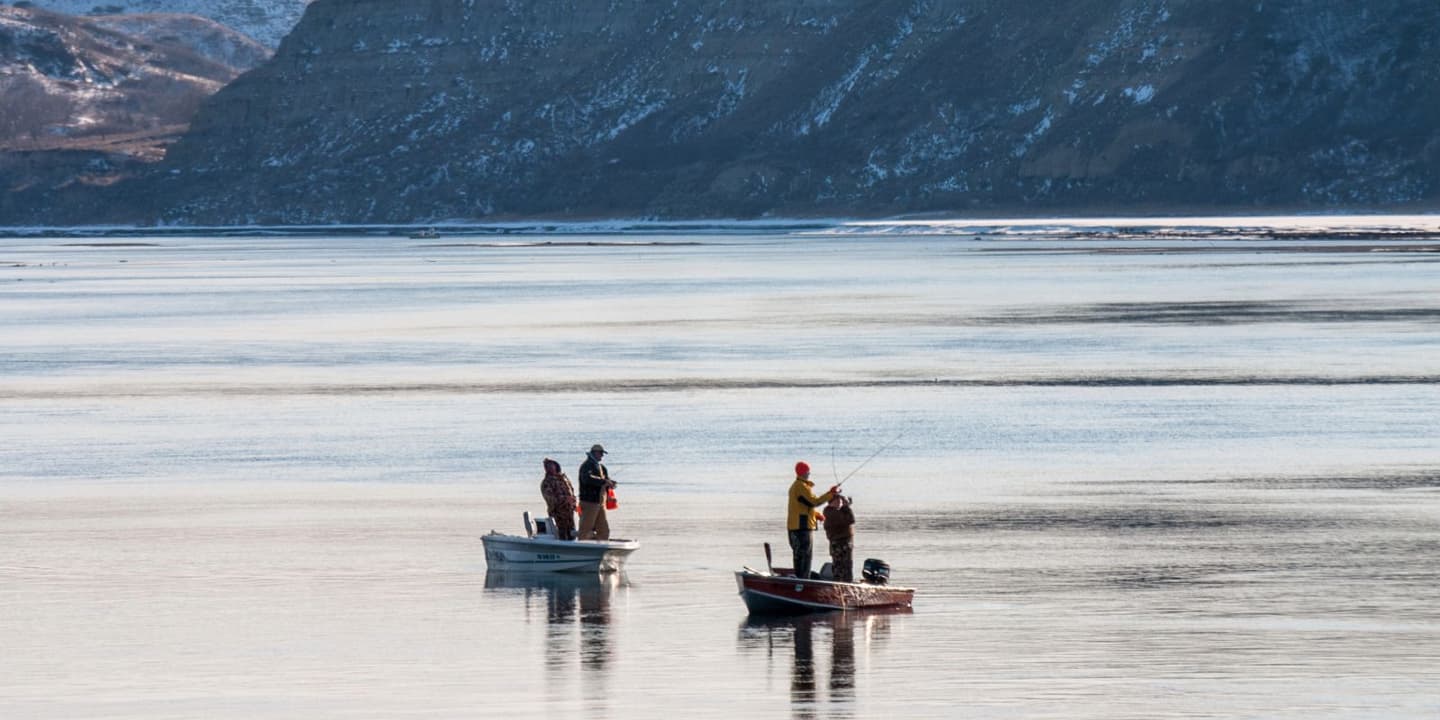 Two small boats with people fishing on a calm lake, set against a backdrop of snow-covered mountains in North Dakota