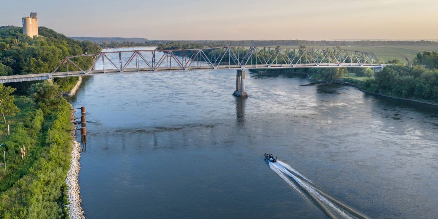 A motorboat travels along a wide river, reminiscent of scenes covered by Nebraska boat insurance, passing under a large metal truss bridge with a tall grain silo visible on the wooded bank.