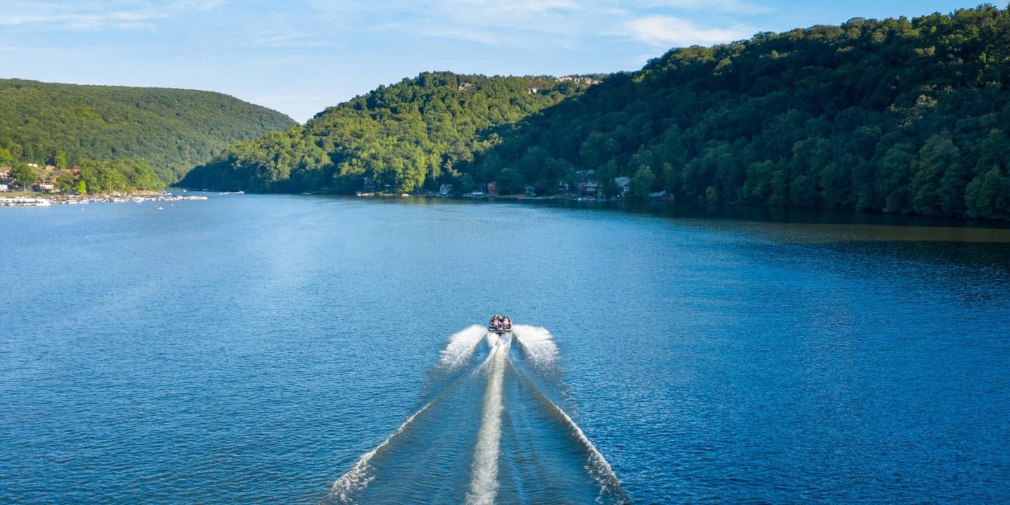 A motorboat leaves a wake while traveling through a calm lake surrounded by forested hills and waterfront houses in West Virginia