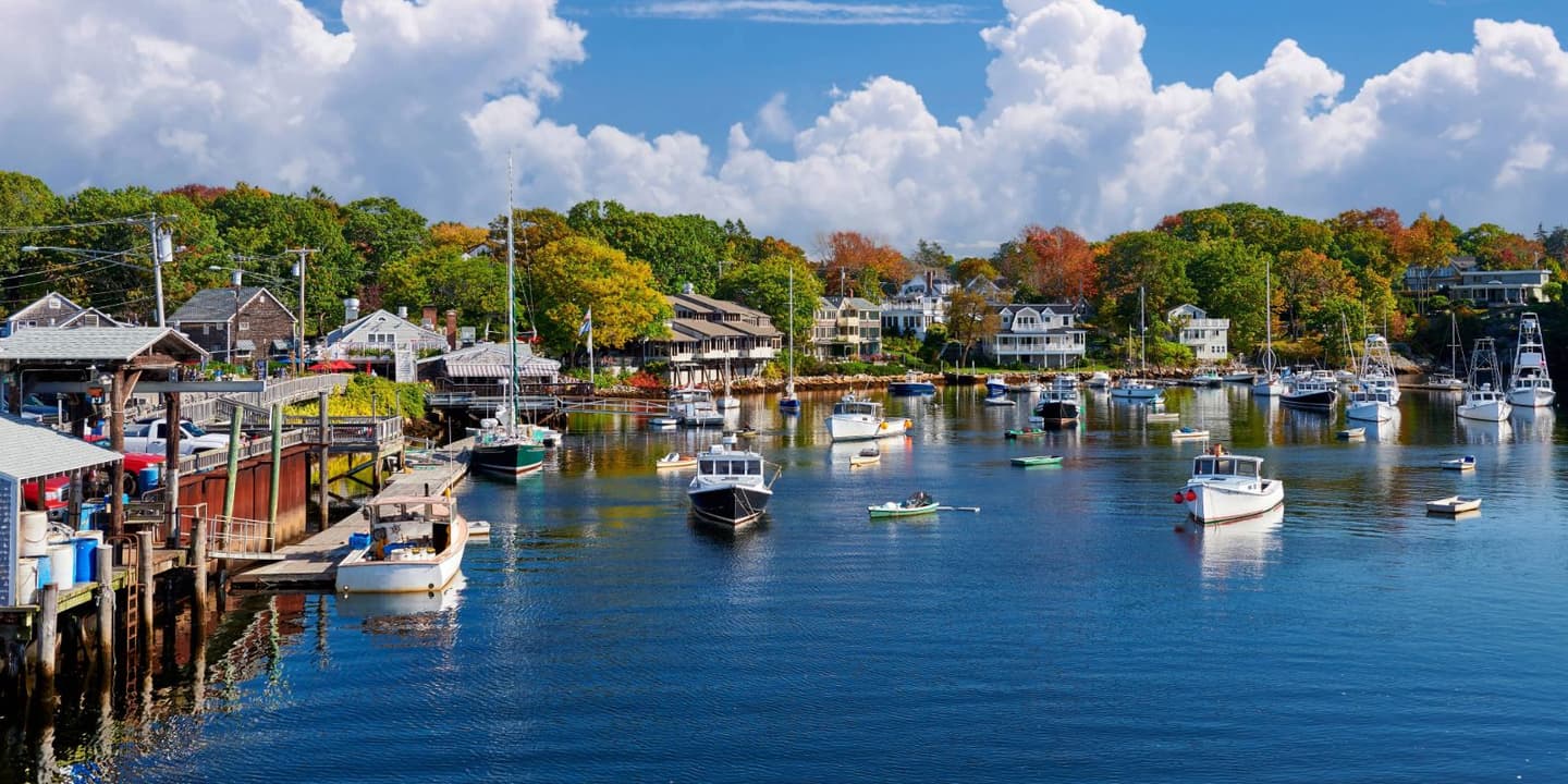 A scenic harbor filled with small boats, often covered by Maine boat insurance, docked before houses nestled among autumn-colored trees under a cloudy blue sky.
