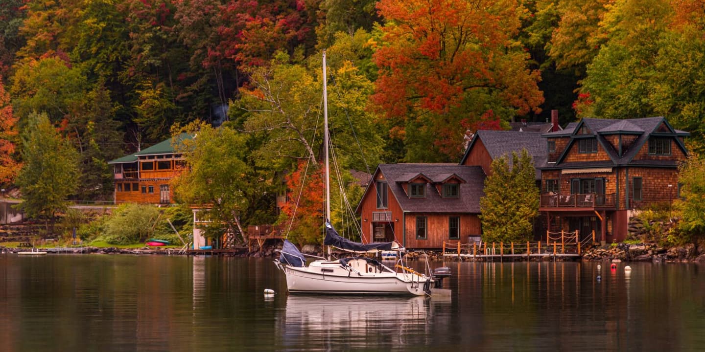 A white sailboat anchored on a calm lake in front of Vermont houses surrounded by autumn trees.