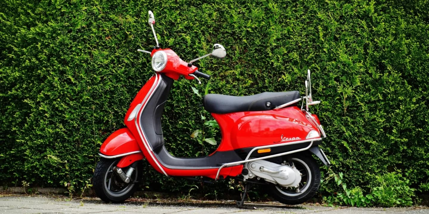Red Vespa scooter with black seat and chrome accents parked in front of a green hedge