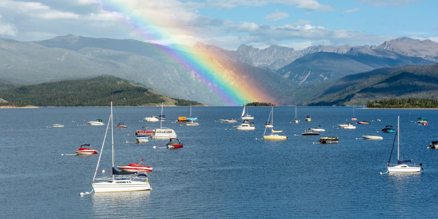 A rainbow arches over a mountain lake filled with numerous sailboats and motorboats with Colorado boat insurance.