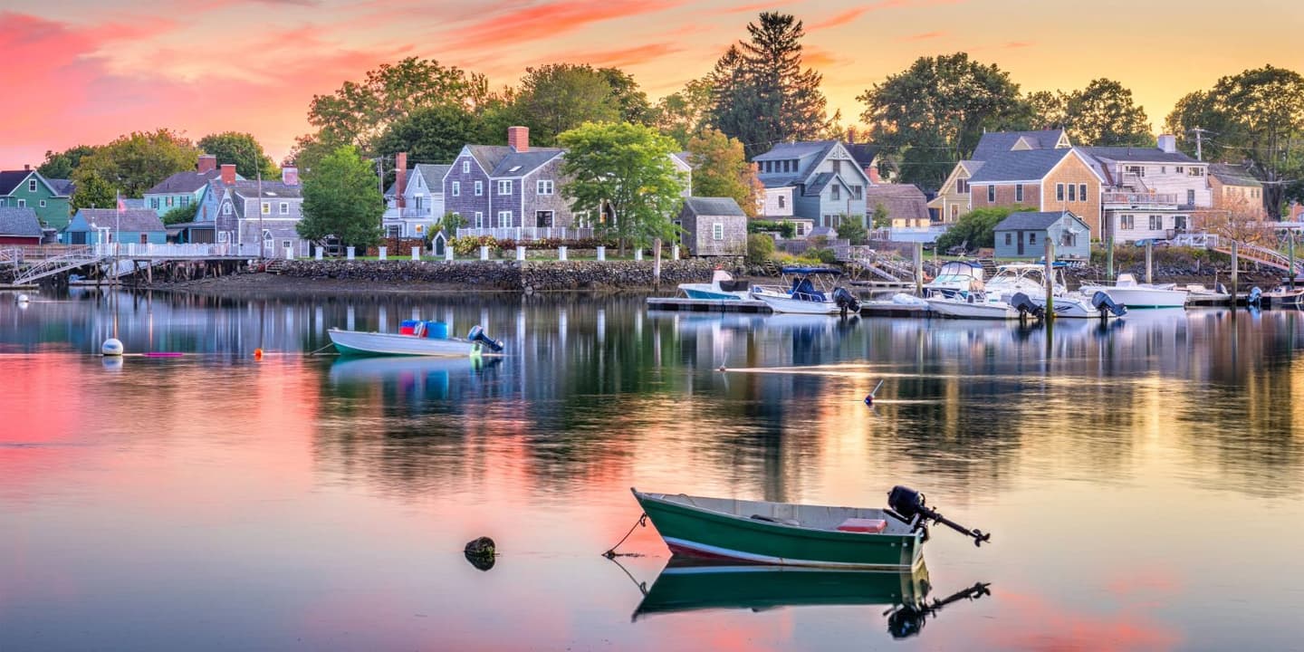 Small boats bob in a calm harbor at sunset, reflecting the pink and orange sky in front of a coastal New England town where many vessels rely on New Hampshire boat insurance.