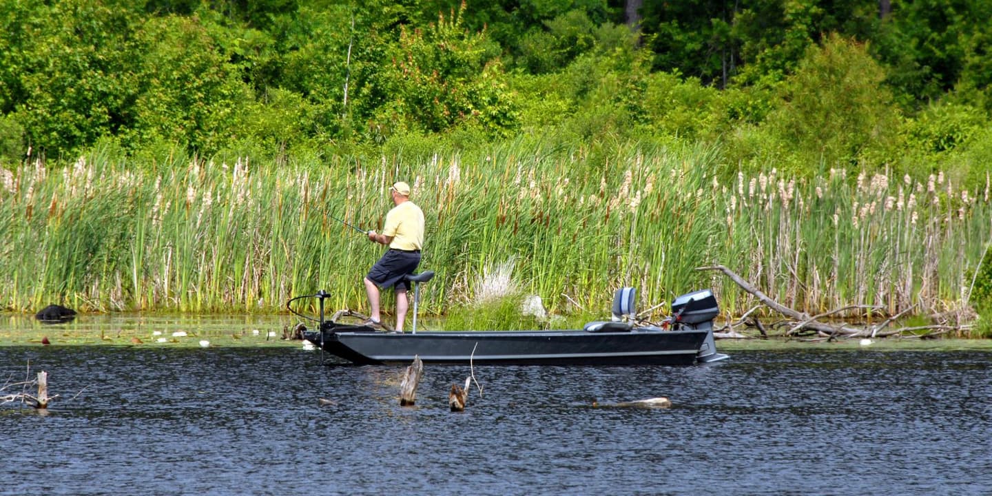 A person in a yellow shirt and cap fishes from a small motorboat near a grassy shoreline, a common sight for those with Arkansas boat insurance.