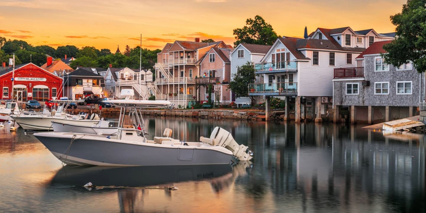 Boats moored in a Massachusetts harbor in front of houses and a red town building at sunset.