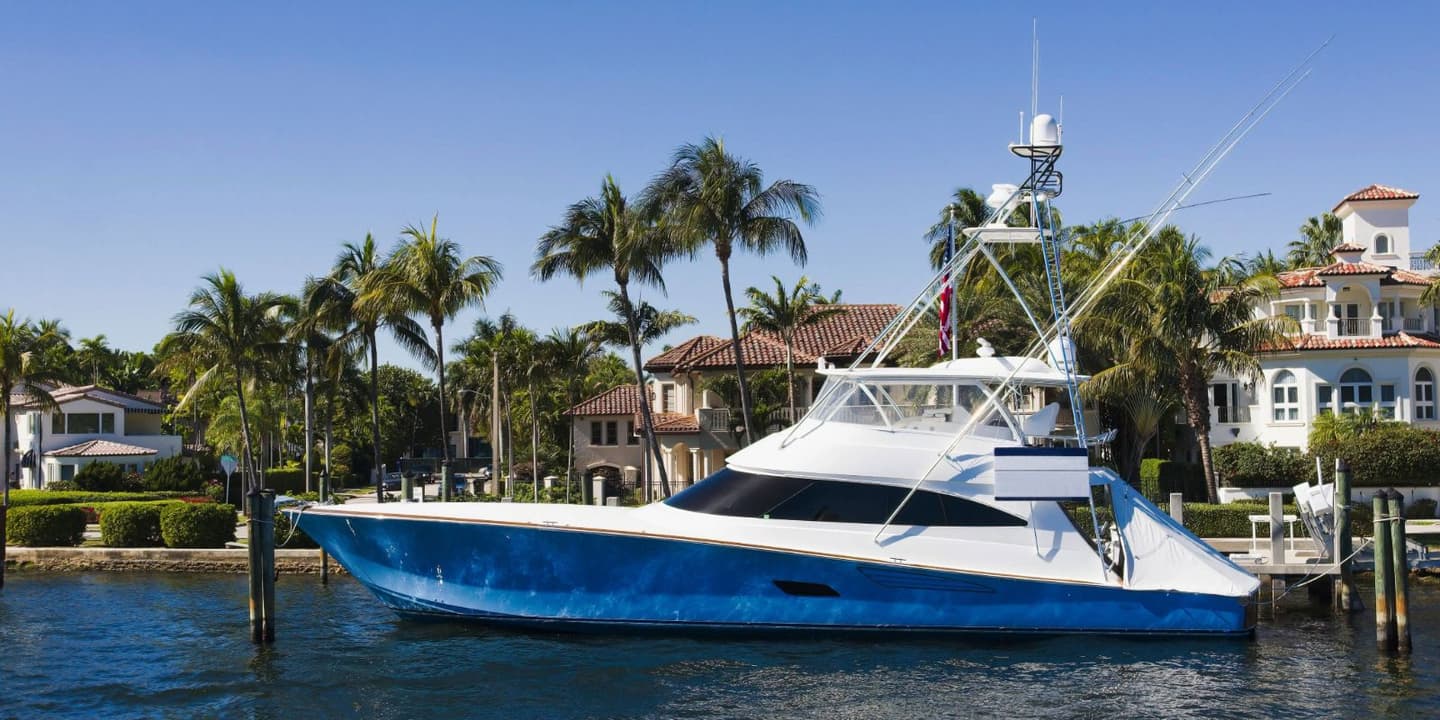 A blue and white motor yacht needing Florida boat insurance moored at a dock in front of waterfront houses and palm trees.