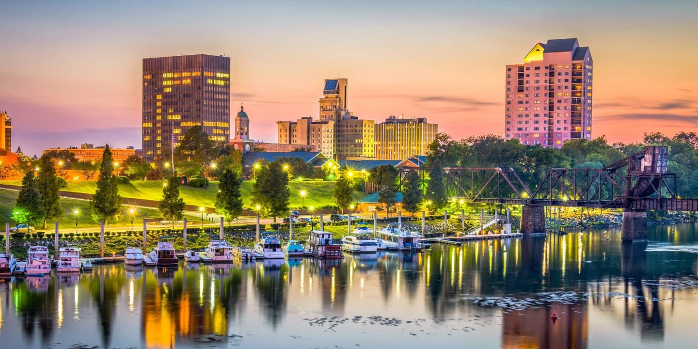 The Augusta, Georgia skyline at sunset, featuring buildings and a railroad bridge reflected in the Savannah River, with a row of boats, perhaps covered by Georgia boat insurance, docked along the riverbank.