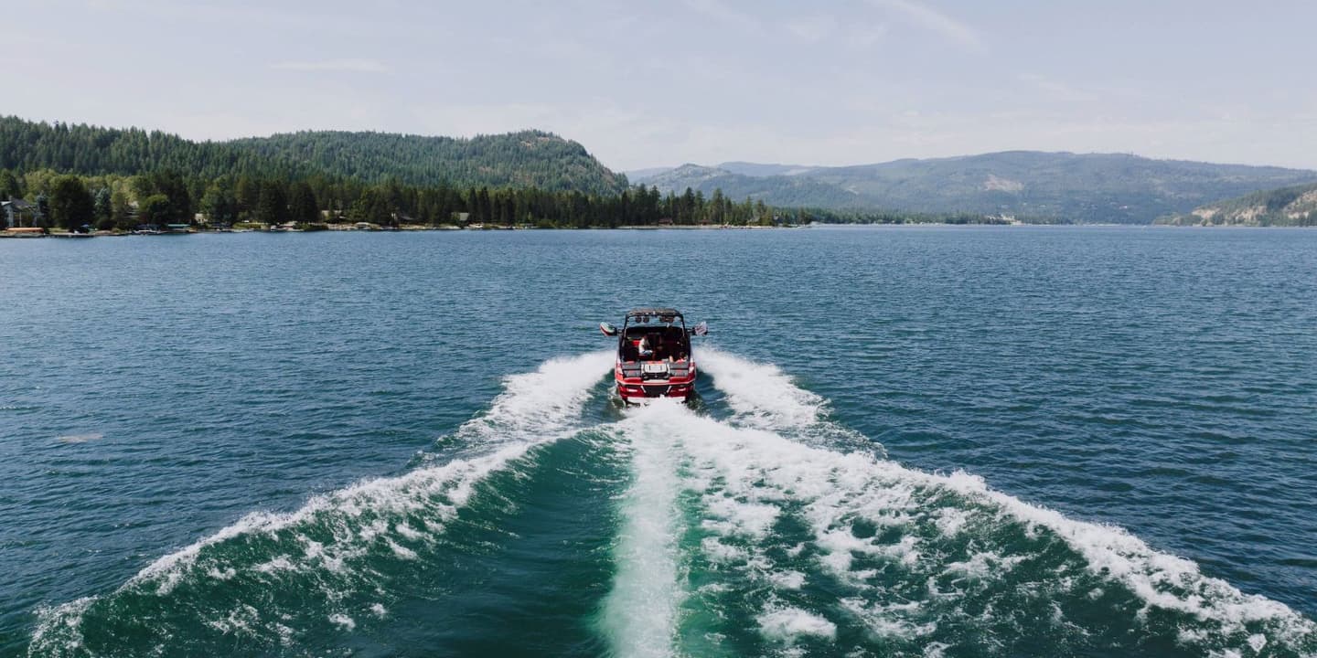 A red motorboat, protected by Idaho boat insurance, moves across a large lake, creating a wide wake toward tree-covered mountains.