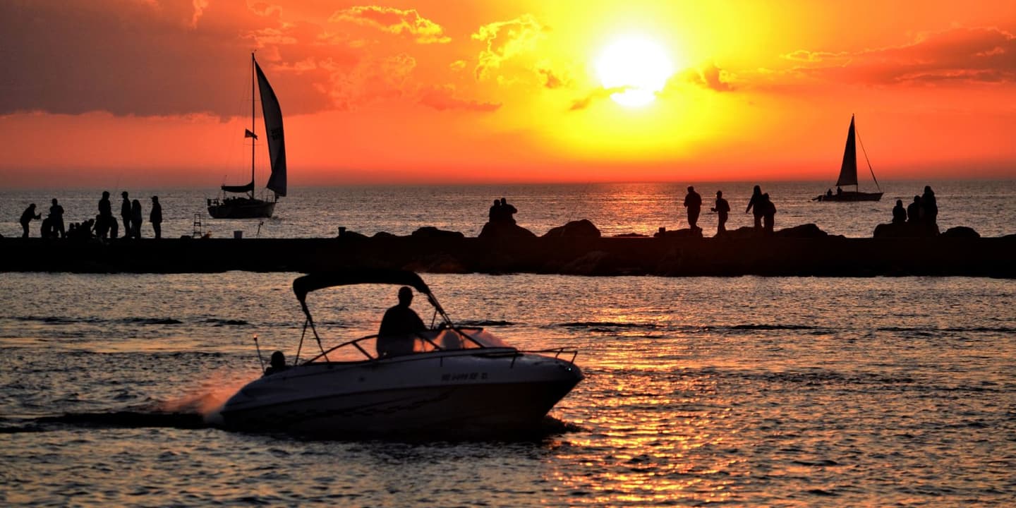 Silhouettes of a motorboat and sailboats on the water against a bright orange sunset, with people standing on a jetty in the distance, evoking the peace of mind found with Michigan boat insurance.