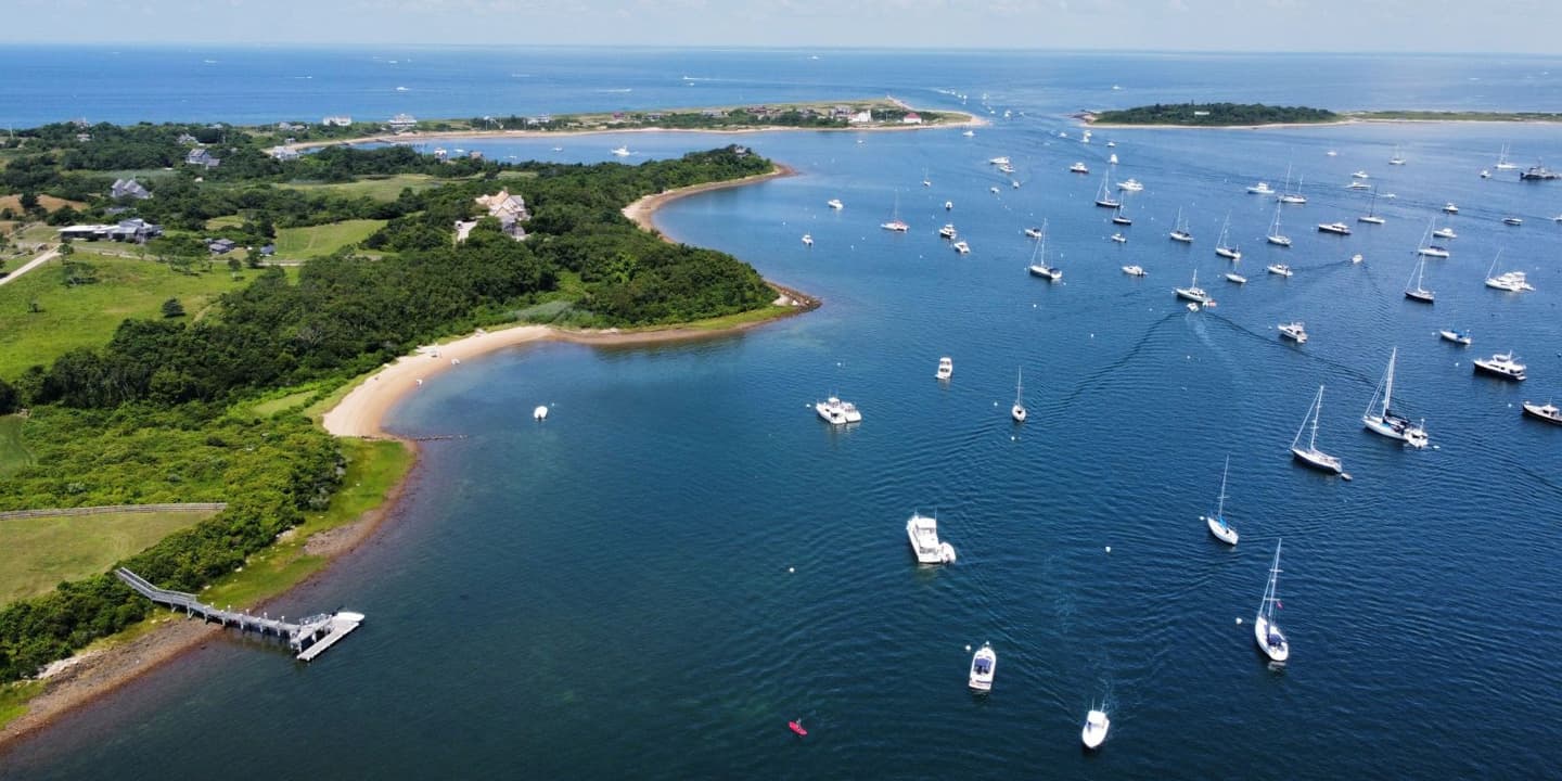 Aerial view of boats anchored in a bay next to a lush, grassy coastline with a wooden pier, a common sight for Rhode Island boat insurance policyholders.