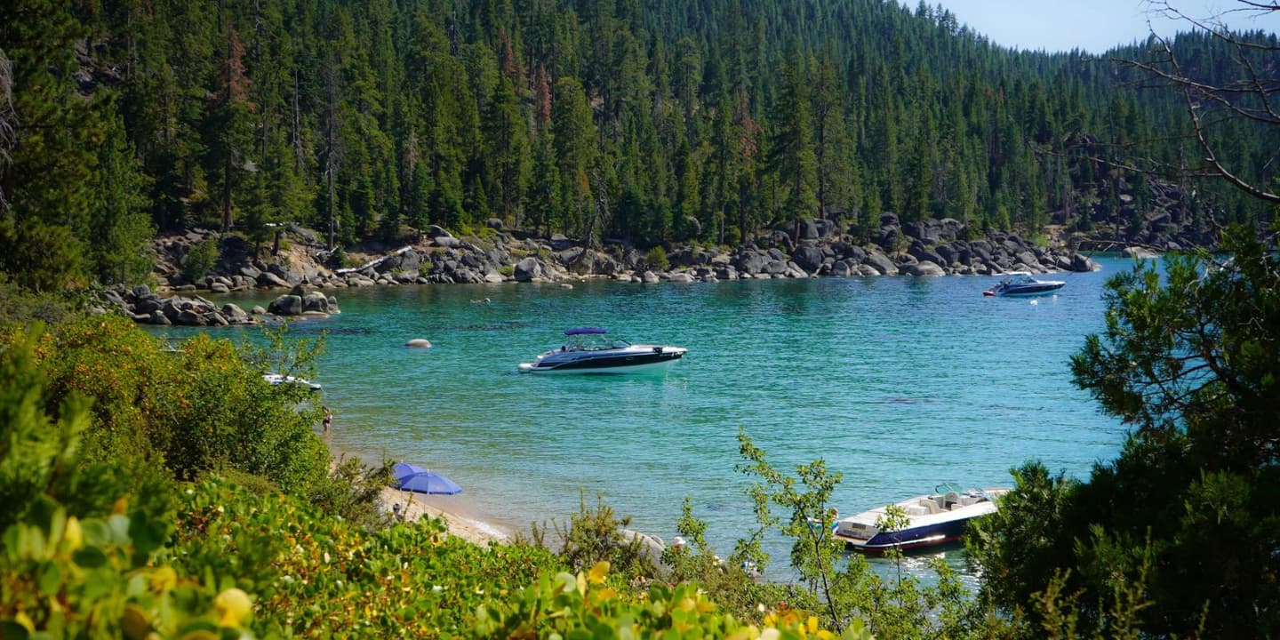 Boats insured by California Boat Insurance rest on the turquoise water of a lake framed by a dense pine forest and a rocky shoreline.