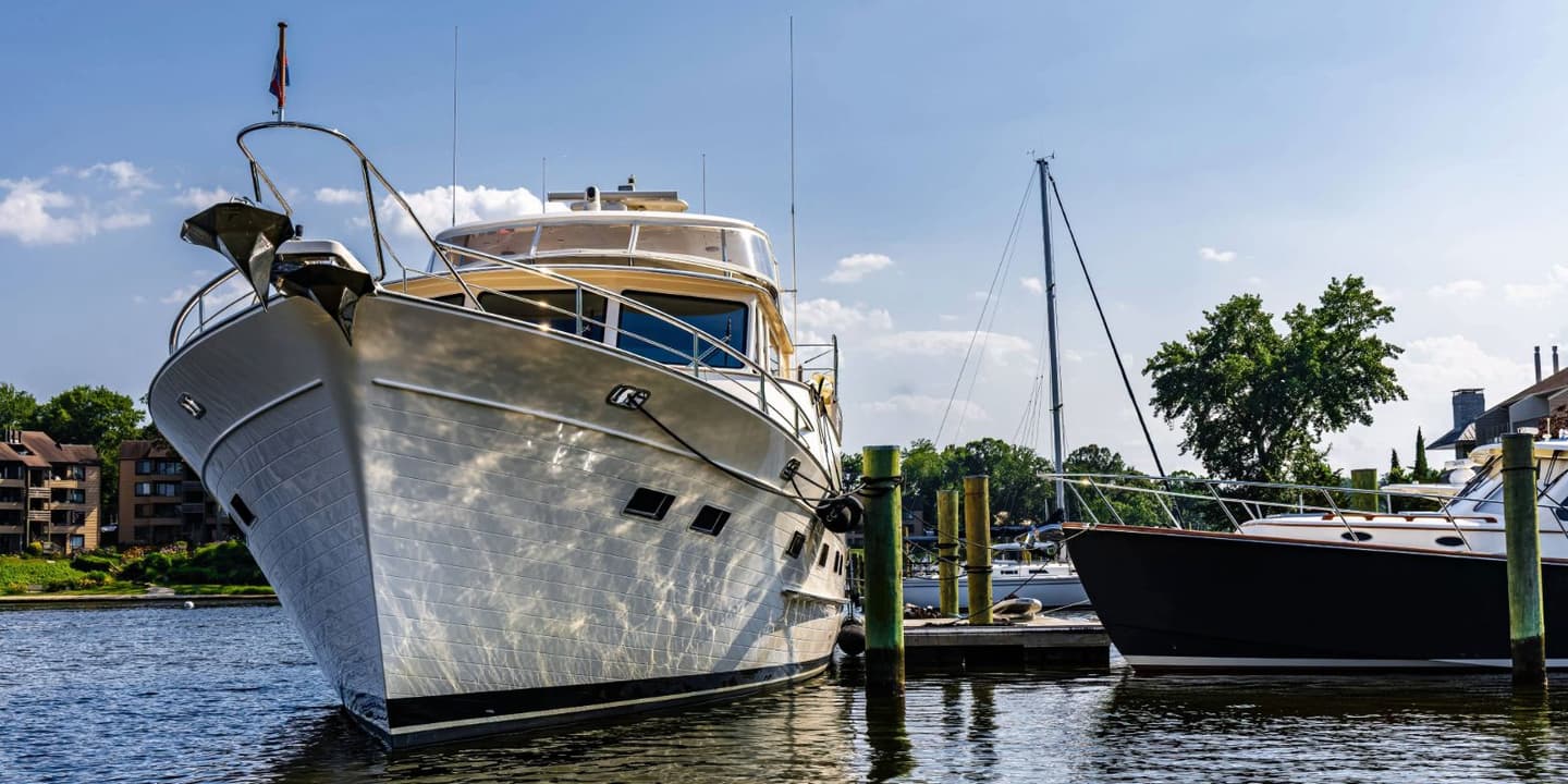 A large white motor yacht and a smaller dark-hulled boat moored at a wooden dock on a sunny day, the type of vessels often covered by Maryland boat insurance.