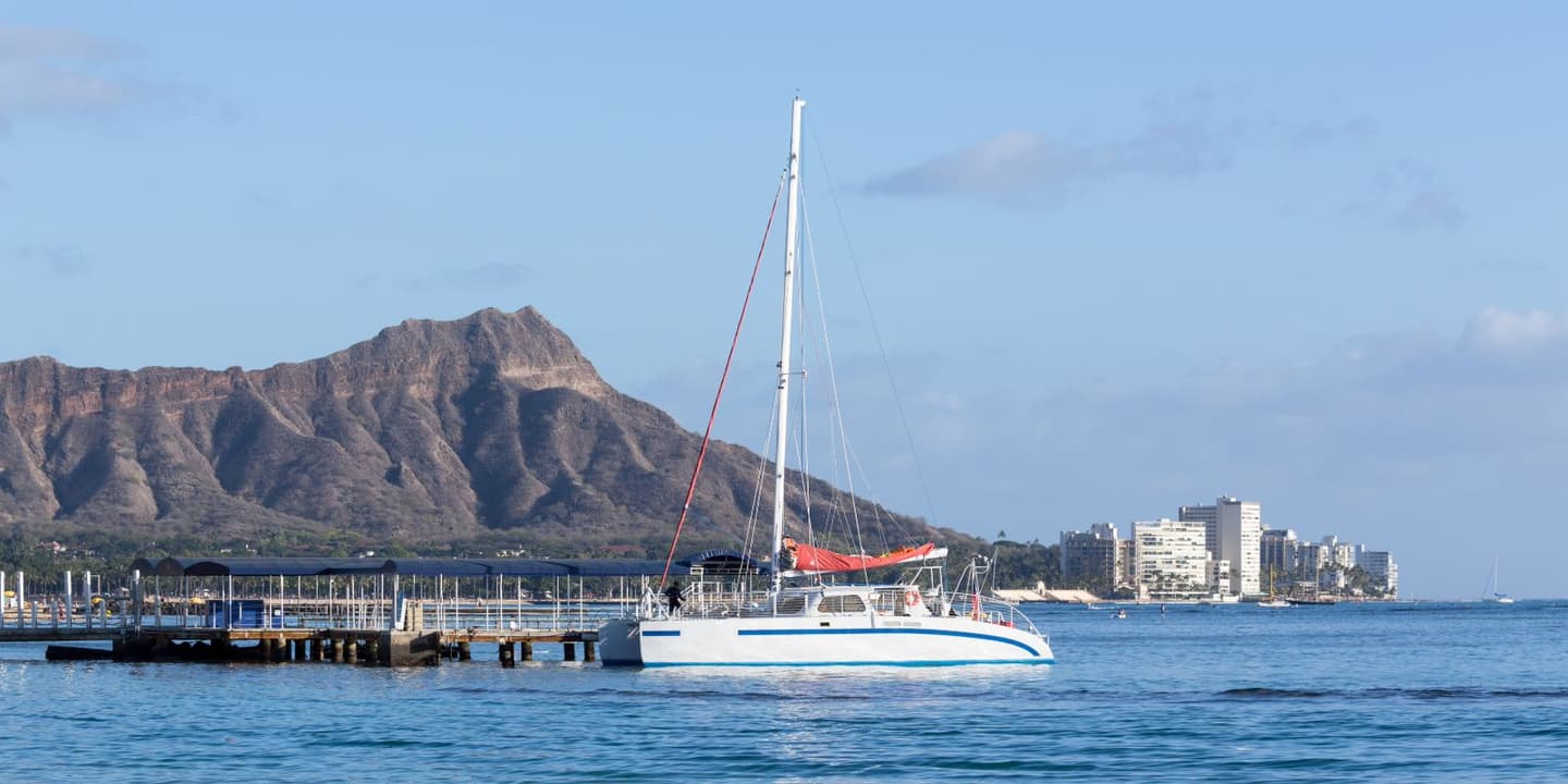 A white catamaran requiring Hawaii boat insurance docked at a pier in front of Diamond Head crater, with a cityscape visible in the distance.