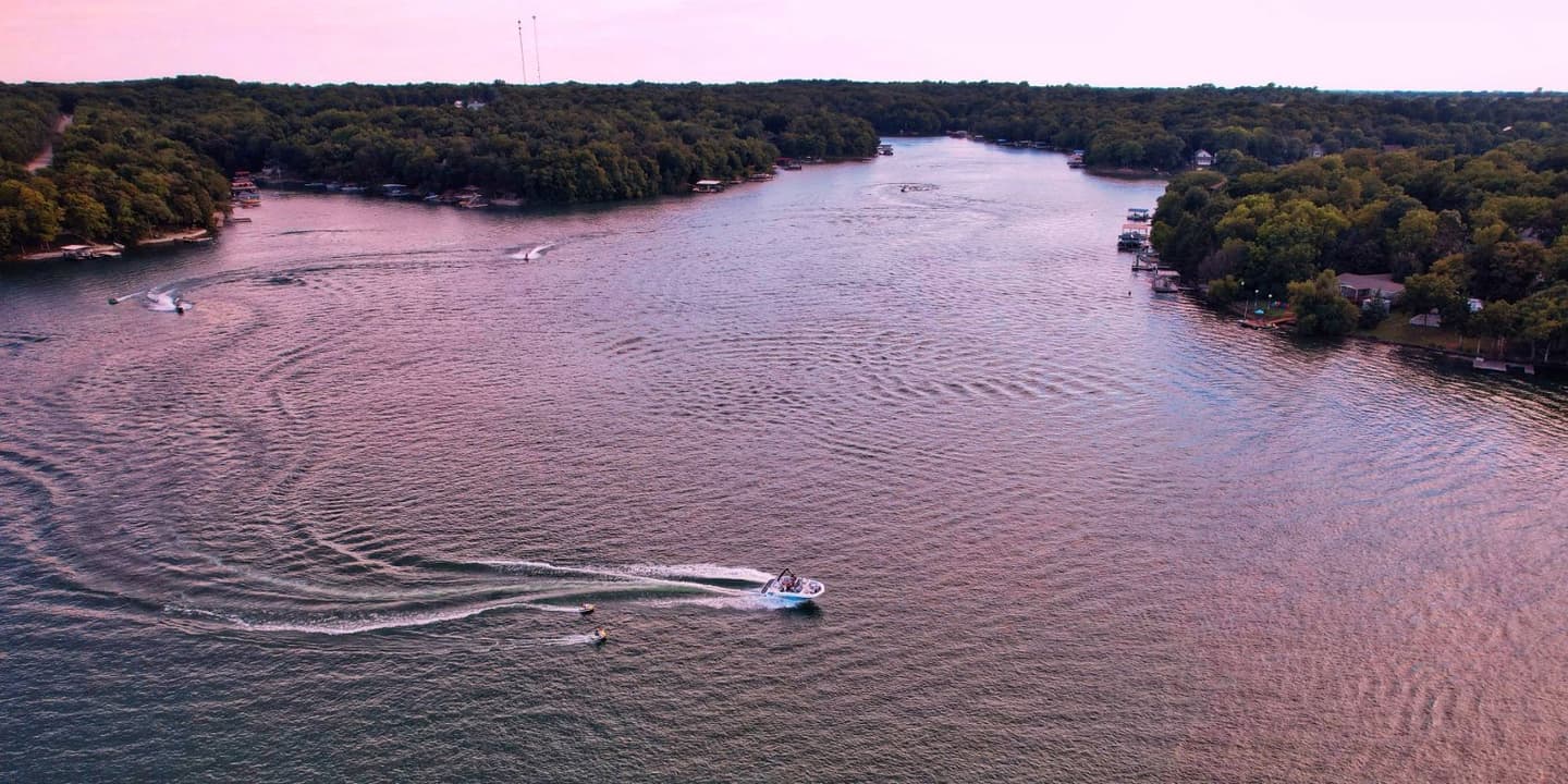 Aerial view of a motorboat, protected by Kansas boat insurance, pulling a person on a tube across a large, tree-lined lake at sunset.