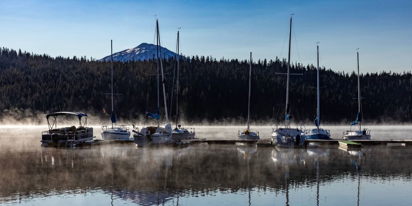 Sailboats and a pontoon boat, like those protected by Oregon boat insurance, docked on a misty lake with a snow-capped mountain in the distance.