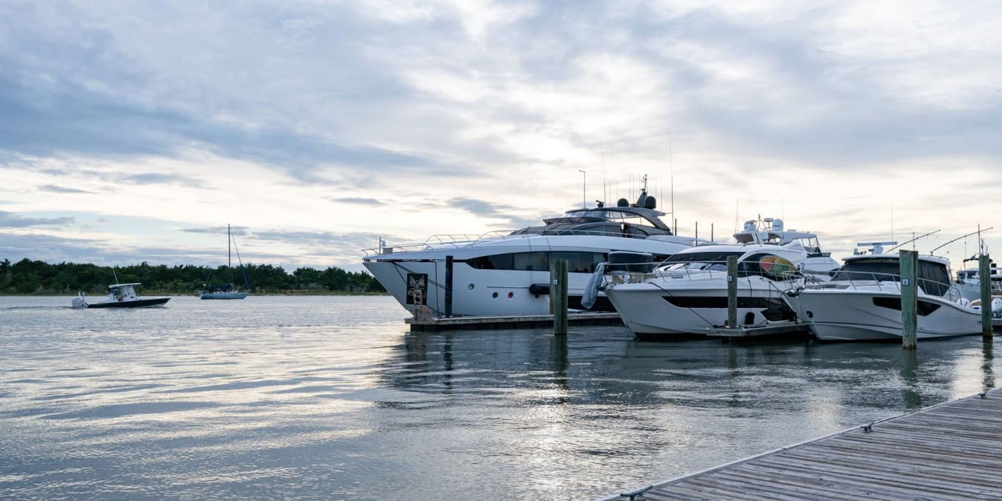Three luxury yachts, the kind often protected by North Carolina boat insurance, docked at a pier at dusk, with two smaller boats sailing on the water in the distance.