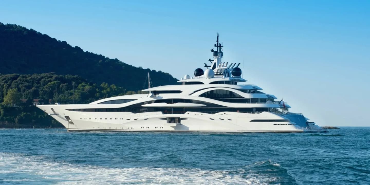 Large white luxury motor yacht with multiple decks cruising near a wooded coastline