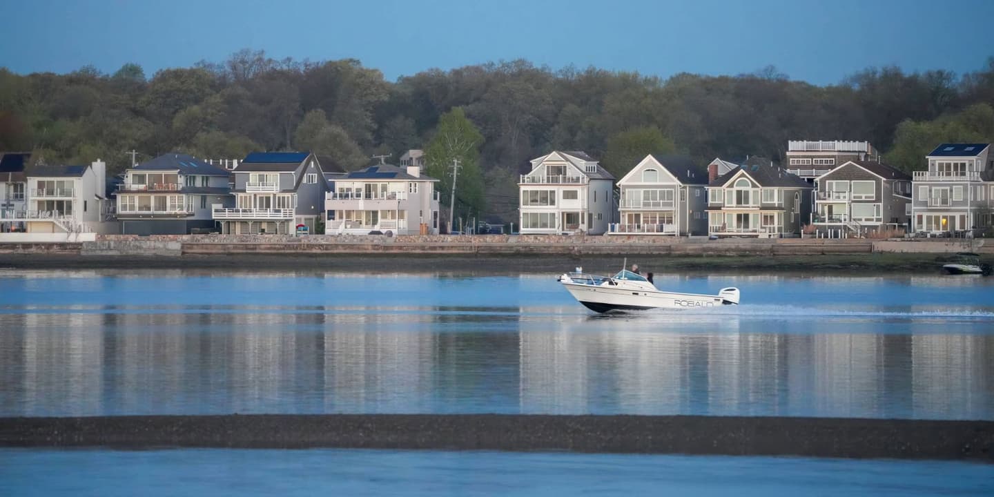 A white motorboat, the type often covered by Connecticut boat insurance, cruises on calm water in front of a row of houses lining the shore.