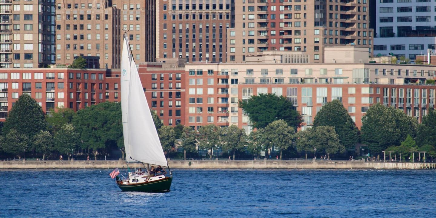 A sailboat with a white sail and an American flag on the stern cruises on a river in front of city apartment buildings and trees, a common scene for New Jersey boat insurance coverage.