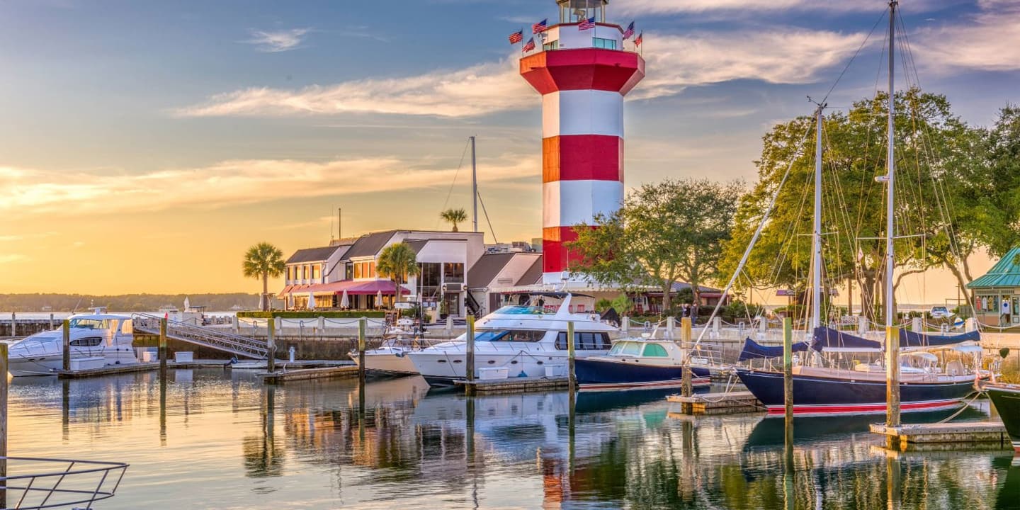 The red and white striped Harbour Town Lighthouse stands above a marina filled with boats at sunset,