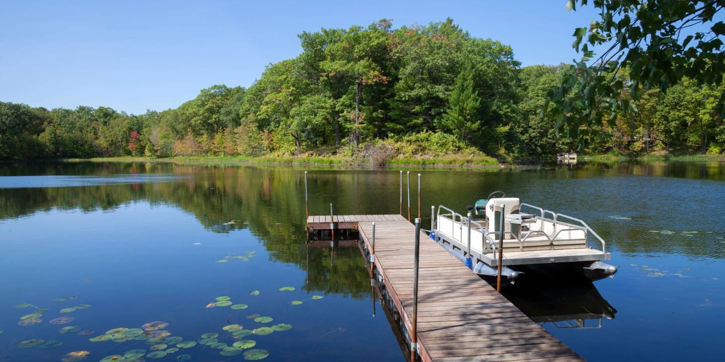 A pontoon boat docked at a wooden pier on a calm, tree-lined lake, a common sight for those with Minnesota boat insurance.
