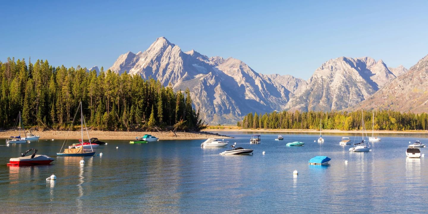 Many motorboats and sailboats with Wyoming boat insurance anchored on a calm lake in front of a dense pine forest and jagged, snow-dusted mountains.