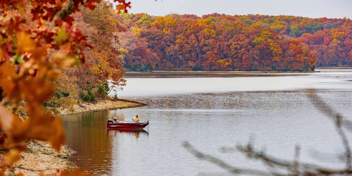 A person in a yellow jacket sits in a small red motorboat on a calm lake surrounded by autumn trees.
