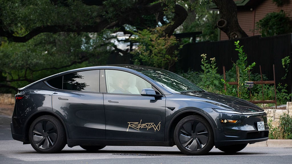 A Tesla Model Y Robotaxi in stealth grey with a gold 'Robotaxi' script decal, rolling through a leafy Austin, Texas neighborhood.